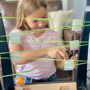 May include: A child plays a game with a ping pong ball and elastic bands stretched across a black chair. The elastic bands are attached to white cards with scripture references written on them, including "Alma 36:17-24", "Mosiah 7:14", and "Alma 30:38".