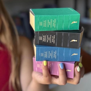 Puede incluir: Una pila de cuatro libros en verde, negro, azul y rosa. Cada libro tiene letras doradas y un pequeño emblema dorado. Los títulos incluyen "Holy Bible", "Book of Mormon", "Doctrine and Covenants" y "Pearl of Great Price".