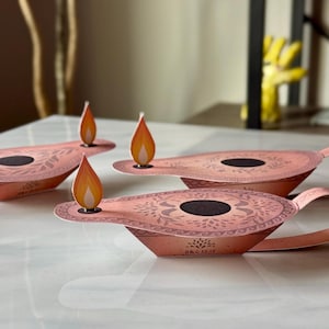 May include: Three paper diya oil lamps with orange flames. The lamps are pink with intricate floral designs and black accents. The lamps are on a white surface.