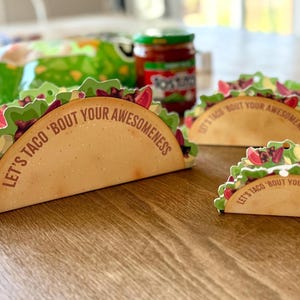 以下が含まれることがあります： 「LET'S TACO 'BOUT YOUR AWESOMENESS」の文字が入った、タコス型の装飾カードが3枚。カードはタン色のベースに、レタス、トマト、その他の具材がカラフルにトッピングされています。背景にはチップスの袋とサルサの瓶があります。