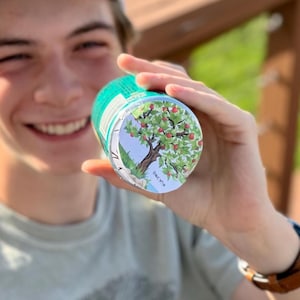 May include: A cylindrical container with a teal background and a colorful illustration of an apple tree with red apples. The container is held in a person's hand.