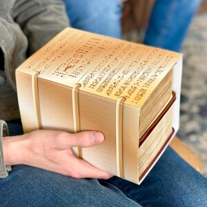 Könnte beinhalten: Eine Person hält eine große, rechteckige, braune Schachtel mit einem weißen Etikett. Das Etikett ist mit Text bedeckt, der schwer zu lesen ist. Die Schachtel scheint aus Holz oder Pappe zu sein.