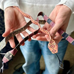 Op de afbeelding: Drie origami-slangen gemaakt van papier met verschillende patronen. De slangen zijn rood, zwart en wit met geometrische patronen. De slangen worden in de handen van een persoon gehouden.