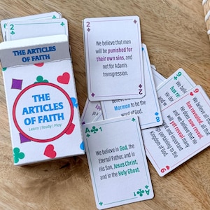 Puede incluir: Una baraja de cartas titulada "The Articles of Faith" con texto en cada carta. La caja es blanca con un círculo rojo y las palabras "The Articles of Faith". Las cartas están sobre una superficie de madera.