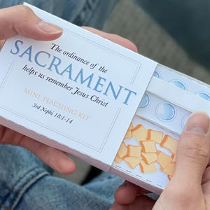 May include: A white box with the text "The ordinance of the SACRAMENT helps us remember Jesus Christ MINI TEACHING KIT 3rd Nephi 18:1-14" printed on it. The box contains small blue and white circles and small yellow squares.