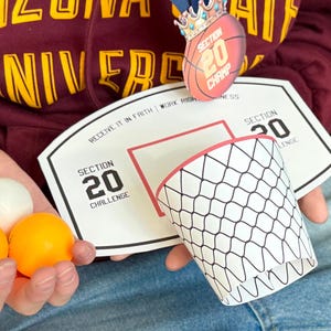 Puede incluir: Un recorte de aro de baloncesto blanco y rojo con el texto "Section 20 Challenge" y "Section 20 Champ" en él. Tres pelotas de tenis de mesa se sostienen en una mano.