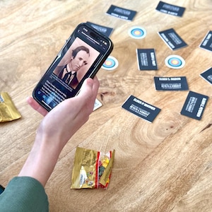 May include: A person is holding a smartphone displaying a photo of a man with a beard and a black suit. The phone is on a wooden table with several business cards and small round stickers.