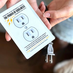 Puede incluir: Una cubierta decorativa blanca para tomacorrientes con dos enchufes, con el texto "THE POWER OF GODLINESS IS MANIFEST. D&C 84:20". Un enchufe negro está conectado. La cubierta es sostenida por una persona.