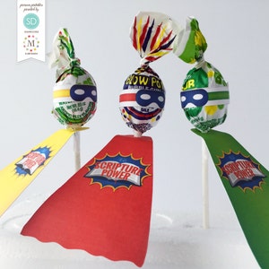 May include: Three lollipops with red, yellow, and green superhero capes. The lollipops are wrapped in colorful foil and have a blue and black eye mask design. The capes have the words "Scripture Power" printed on them.