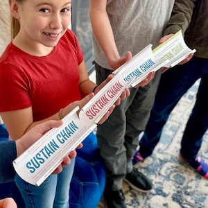 May include: Four people hold up white tubes with the words "Sustain Chain" printed on them in blue and red letters. The tubes are connected together to form a chain.