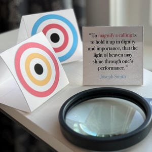 May include: Two white triangular targets with concentric circles in red, yellow and black. A magnifying glass and a white card with a quote by Joseph Smith: "To magnify a calling is to hold it up in dignity and importance, that the light of heaven may shine through one's performance."