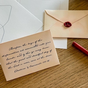 May include: A collection of stationery items on a wooden surface. Includes a light brown envelope with a red wax seal, a red pencil, and a light brown card with handwritten text from Doctrine & Covenants 110:16. Also, there are several white envelopes.