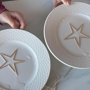 May include: Two white plates with a woven pattern on the rim. Each plate has a wooden star shape glued to the center. The stars are made of thin wooden sticks.
