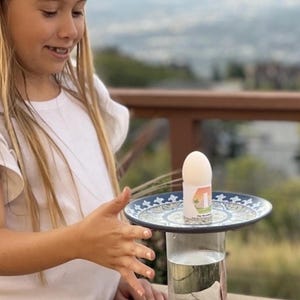 Op de afbeelding: Een wit ei ligt op een blauw en wit keramiek bord, dat op een heldere glazen pot is gebalanceerd. De pot is gevuld met water. Het ei is versierd met een kleurrijke sticker met de tekst "The Big Brunch".