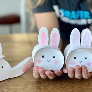 May include: Three white paper bunny boxes with pink ears and pink cheeks. The boxes are decorated with black eyes and a black nose.