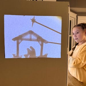 May include: A cardboard shadow box displaying a nativity scene silhouette. The scene includes a stable, figures, and a star. A young person in a yellow sweater is looking at the shadow box.