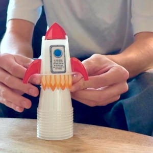 May include: A toy rocket made from a white plastic cup, featuring red fins and a red cone-shaped top. The rocket has a blue circle and a small rectangular label on the front, with orange flames at the base.