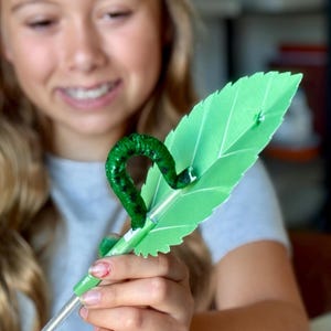 May include: A green paper leaf with a green caterpillar decoration. The caterpillar is a vibrant green with darker green spots. The leaf has a detailed vein pattern and is held by a silver pen.