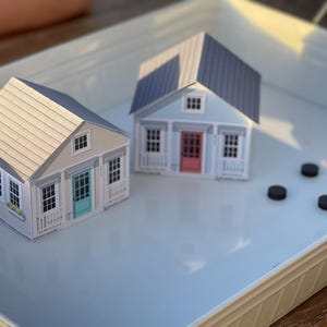 以下が含まれることがあります： 白いトレイの上に、ターコイズ色のドアが付いた灰色のミニチュアペーパーハウスと、赤いドアが付いた青いミニチュアペーパーハウスが2つ置かれています。トレイには黒い丸いものが4つあります。