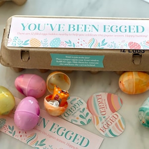 May include: A brown cardboard egg carton with a label that says "You've Been Egged!" There are 12 filled eggs hidden nearby to help you celebrate this joyous season. Happy hunting!" The carton is surrounded by colorful plastic eggs and stickers that say "We've Been Egged!"