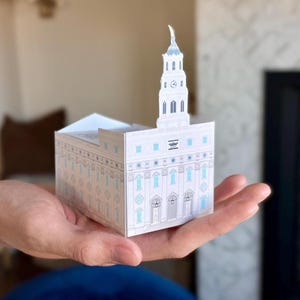 May include: A white and light blue paper model of a building with a tall steeple, held in a hand. The model has detailed architectural features, including windows and decorative elements. The building is a religious structure.