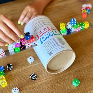 May include: A tabletop game with colorful dice and a cylindrical container. The dice are arranged on a ruler and scattered on a wooden surface. The container has the text "TEETER TOPPLE" printed on it.