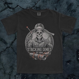 Puede incluir: Una camiseta gris carbón de manga corta con el gráfico de un esqueleto con máscara de soldar, sosteniendo un soplete y un martillo de soldador. El texto STACKING DIMES EST 2026 aparece sobre dos tubos de metal soldados con detalles naranjas.