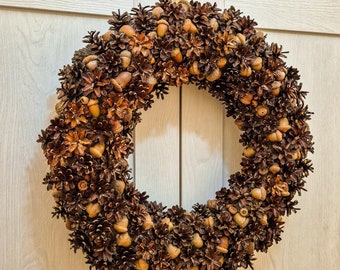 May include: A brown and beige autumnal wreath made of pine cones and acorns. The wreath is hanging on a white wooden door.