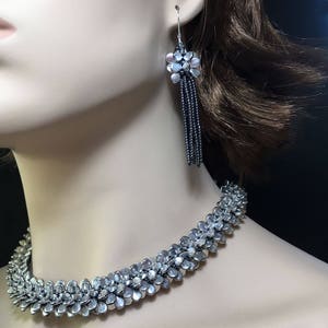 May include: A silver choker necklace and matching dangle earrings. The necklace is made of small, silver beads that are arranged in a flower-like pattern. The earrings are also made of silver beads and have a tassel-like design.