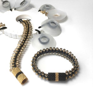 May include: Two beaded bracelets, one gold and black, the other black and brown, with a gold and black end cap. The bracelets are on a white surface.