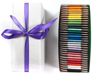 May include: A white gift box tied with a purple satin ribbon next to a box of colorful embroidery floss. The floss is arranged in a rainbow of colors, from yellow to red, with a variety of blues and greens.