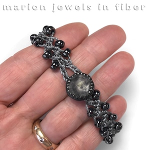 May include: A handmade bracelet featuring a central, dark gray, circular element with a textured surface, connected by gray woven threads. The bracelet is adorned with small, black beads. The text "marion jewels in fiber" is at the top.
