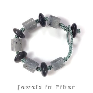 Pode incluir: Um bracelete de contas cinzas e pretas com um cordão verde. As contas são esculpidas com desenhos intrincados. O bracelete é amarrado com um nó.