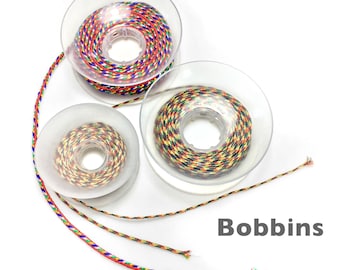 May include: Three clear plastic spools of colorful braided cord. The cord is a mix of red, blue, yellow, and green. The word "Bobbins" is printed in white. The text "marion jewels in fiber" is printed at the top.