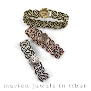 May include: Three black braided bracelets with different colored metal clasps. The clasps are a gold heart, a copper dragonfly, and a silver heart. The bracelets are made with a woven design and are called "marion jewels in fiber".