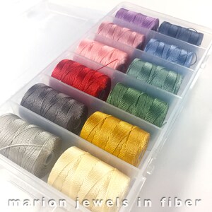 May include: A clear plastic storage box filled with spools of colorful embroidery floss. The floss is arranged in rows, with each row containing a different color. The colors include purple, pink, light pink, red, gray, green, yellow, and cream.