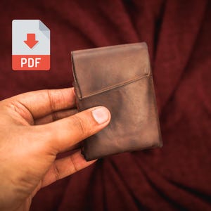 Op de afbeelding: Een bruine leren etui met een klepsluiting. De etui is rechthoekig en lijkt gemaakt te zijn van een getextureerd leermateriaal. Een PDF-pictogram staat in de linkerbovenhoek.