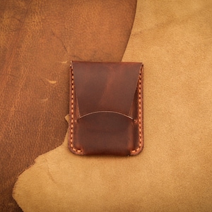 May include: A handcrafted, dark brown leather cardholder with a curved front pocket and a flap closure. The edges are stitched with light brown thread. The cardholder is displayed on a textured, two-tone brown leather surface.