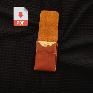 May include: A tan leather card holder with a vertical flap and visible stitching. The interior of the flap shows a rough, suede-like texture, while the exterior is smooth. A single card is partially inserted into the main pocket of the holder.