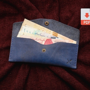 Peut inclure: Un portefeuille en cuir bleu avec une fermeture à bouton-pression, contenant des billets de banque. Le portefeuille présente un design cousu et un petit logo. Le fond est un tissu rouge foncé. Une icône PDF est en haut à droite.