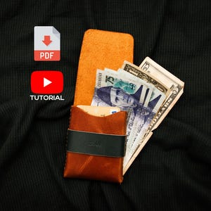 May include: A brown leather card holder wallet with a black elastic band across the front. The wallet is open and contains several paper banknotes, including a seventy-five denomination bill and a five dollar bill, along with a credit card.