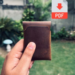 May include: A brown leather wallet with a minimalist design. The wallet is held in a hand against a blurred green background. The wallet has a simple, rectangular shape with a curved top edge and a pocket on the front. A PDF icon is in the upper right corner.