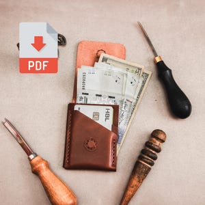 Op de afbeelding: Een bruine lederen kaarthouder met papiergeld en een creditcard, omringd door lederbewerkingsgereedschap, waaronder een metalen priem met een zwart handvat, een houten polijststaaf en een metalen vouwbeen met een licht houten handvat.