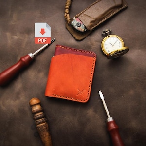 Puede incluir: Primer plano de una cartera de cuero con un esquema de color rojo y naranja. La cartera está sobre una superficie de cuero marrón con una navaja de bolsillo, un reloj de bolsillo y dos herramientas de trabajo del cuero.