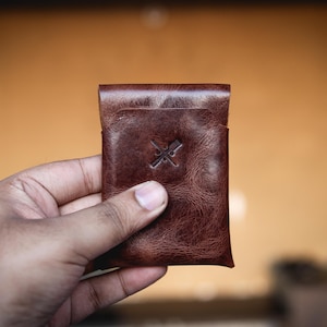 May include: A brown leather cardholder with a textured finish. The cardholder has a debossed logo and a folded top. The leather has a rich, warm tone, and the design is simple and functional.