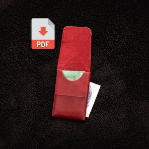 May include: A red leather card holder with a vertical flap and a curved cutout opening. The interior is visible, showing a light green card tucked inside, and a white paper document with a blue graphic pattern is partially visible on the side.