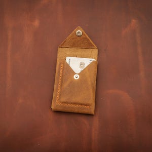 May include: A small, open leather card holder in a warm brown hue, with a triangular flap secured by a silver snap. A white card is partially visible inside. The holder is set against a textured, brown leather background.