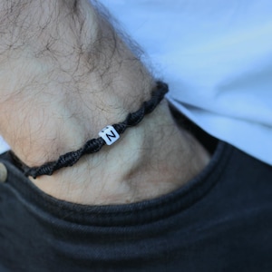 Könnte beinhalten: Ein schwarzes geflochtenes Armband mit einer weißen Perle, auf der der Buchstabe "N" steht.
