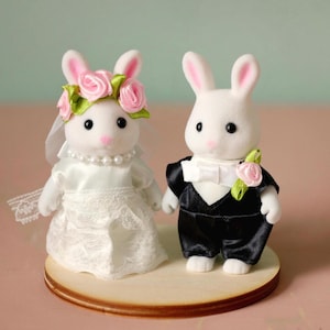 May include: Two white flocked rabbit figurines dressed for a wedding. The left rabbit wears a white satin dress with lace trim, a pearl necklace, and a floral crown with pink roses and green leaves. The right rabbit wears a black tuxedo with a white bow tie and a pink rose boutonniere.