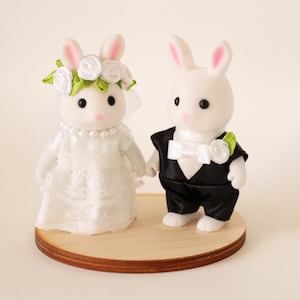 May include: Two white flocked rabbit figurines standing on a circular wooden base. The left rabbit wears a white lace dress, a pearl necklace, and a floral crown. The right rabbit wears a black tuxedo jacket with a white bow tie and a flower.